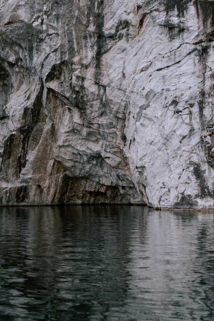 about-02 A stunning rocky cliff with textured granite and marble formations reflected on calm waters.